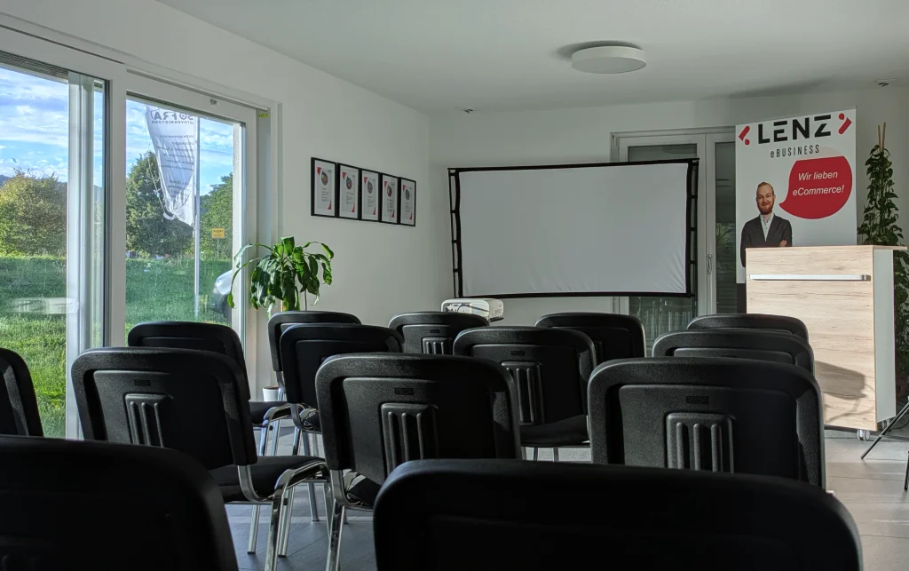 Blick in den Seminarraum Auggen mit Leinwand, holzfarbenem Rednerpult und schwarzen Stühlen. Schöner Blick durch große Fenster nach draußen ins Grüne.